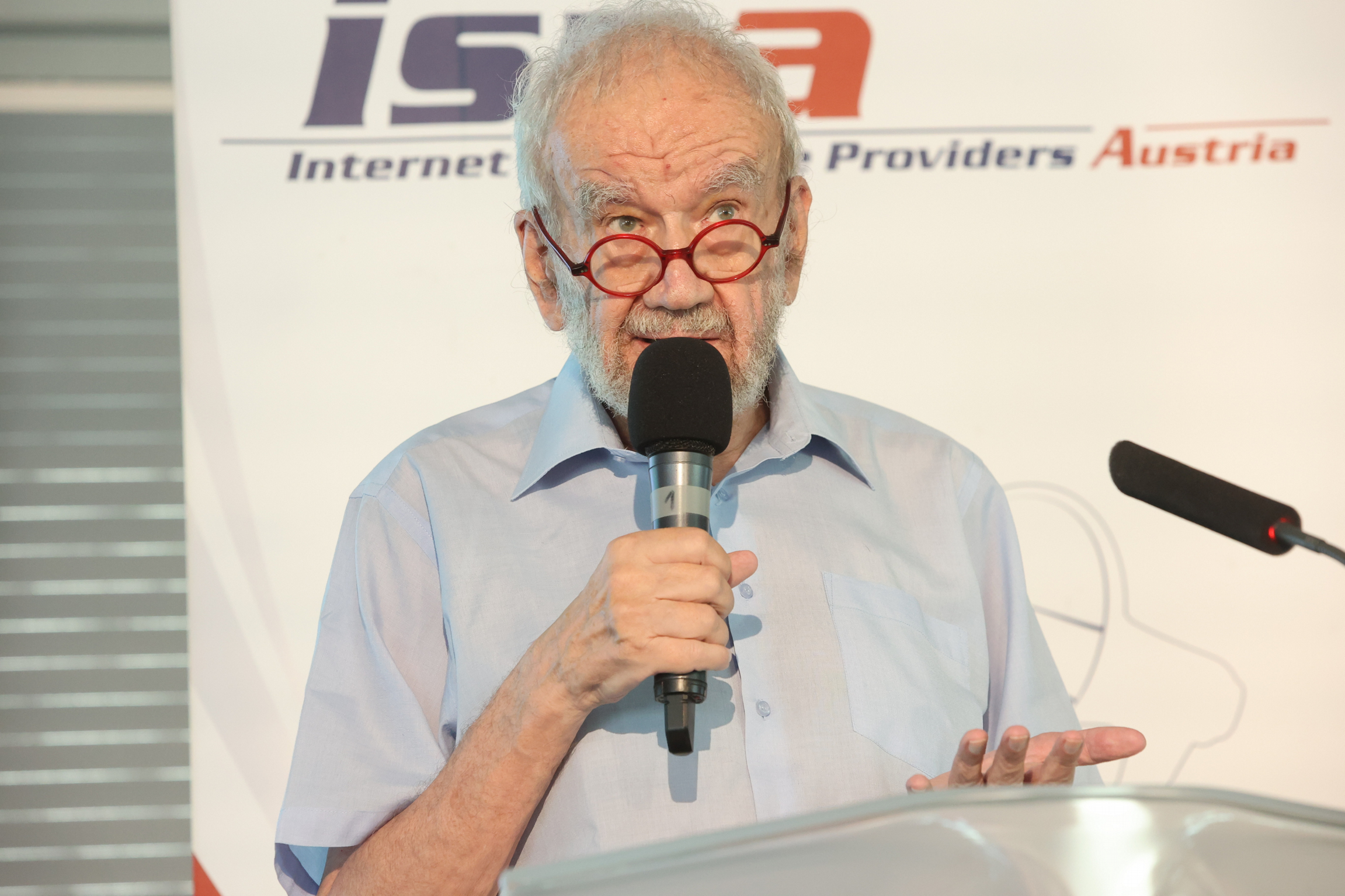 An older gentleman, with a grey beard, grey hair, and red-rimmed spectacles, stands before a sign reading 